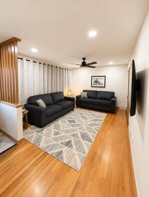A living room with two black sofas, a patterned rug, wooden flooring, a ceiling fan, and a wall-mounted TV. - Midtown Omaha Near Everything! No Cleaning Fee! (Omaha)