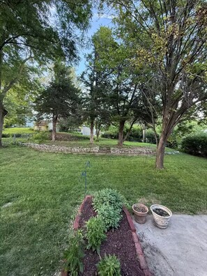 A well-maintained grassy area with a stone pathway, a raised garden bed with plants, and a stone wall in the background. - Midtown Omaha Near Everything! No Cleaning Fee! (Omaha)