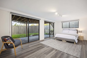 Ein Schlafzimmer mit Bett, Stuhl, einer großen Schiebetür, die in einen Garten führt, und einem Fenster mit Vorhängen.