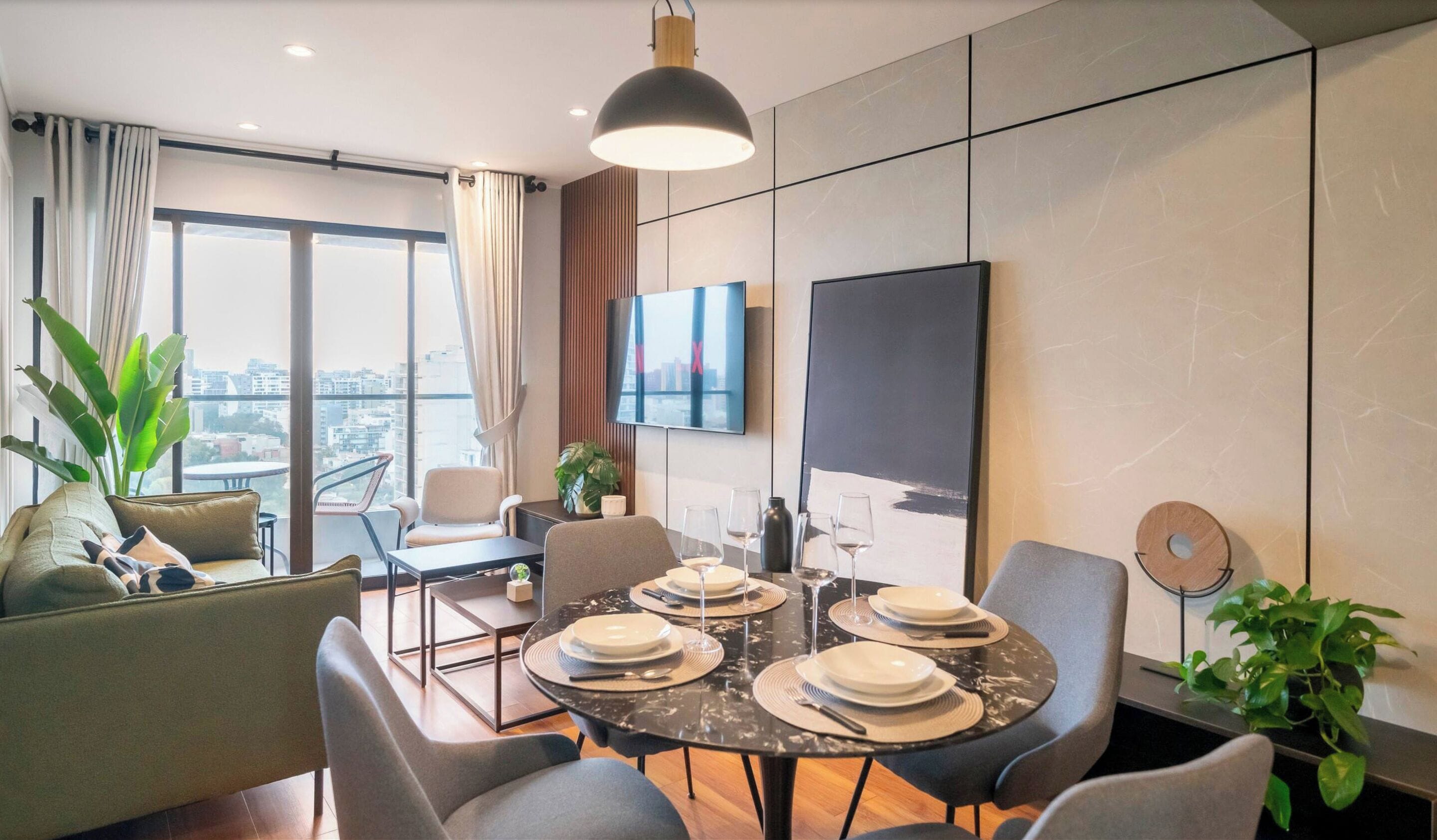 A modern dining area with a round table set for six, a flat-screen TV on the wall, and a city view through large windows.