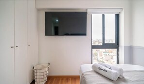 A hotel room with a bed, a TV mounted on the wall, a window with a view of the city, and a laundry basket. - Amazing View 3BR Apartment (Lince)
