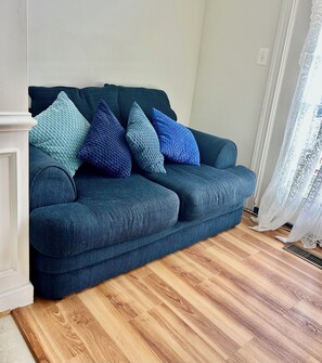 A dark green sofa with multiple cushions in shades of blue and green, positioned against a white wall with a power outlet visible.