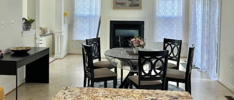 A dining area with a marble table, four chairs, a fireplace, and a large window with patterned curtains.