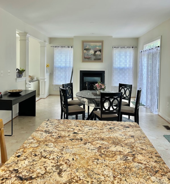 A dining area with a marble table, four chairs, a fireplace, and a large window with patterned curtains.