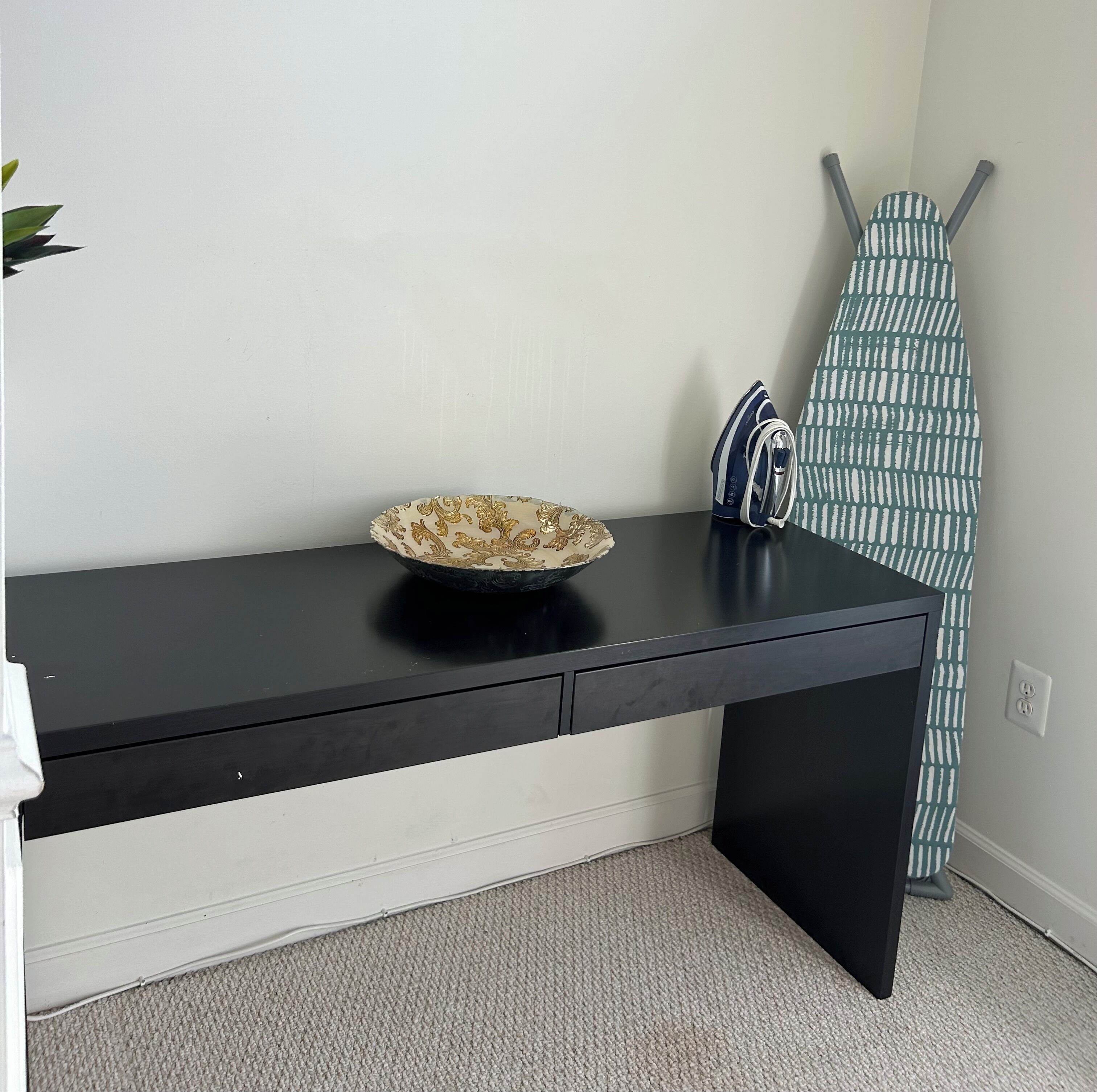 A black desk with a decorative bowl, a blue and white iron, and a patterned ironing board against a white wall.