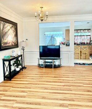 A living room with a flat-screen TV on a stand, a potted plant, a framed picture, and a chandelier.
