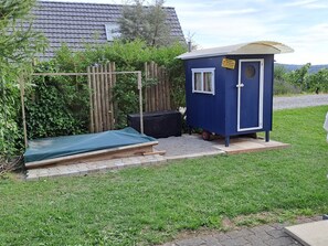 Un cobertizo azul pequeño con puerta y ventana, un banco de madera y una zona cubierta con un lona verde.