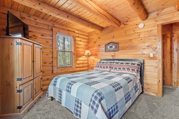 Quarto estilo cabana com teto de madeira, cama com manta estampada, janela com vista para árvores e luminária na parede.