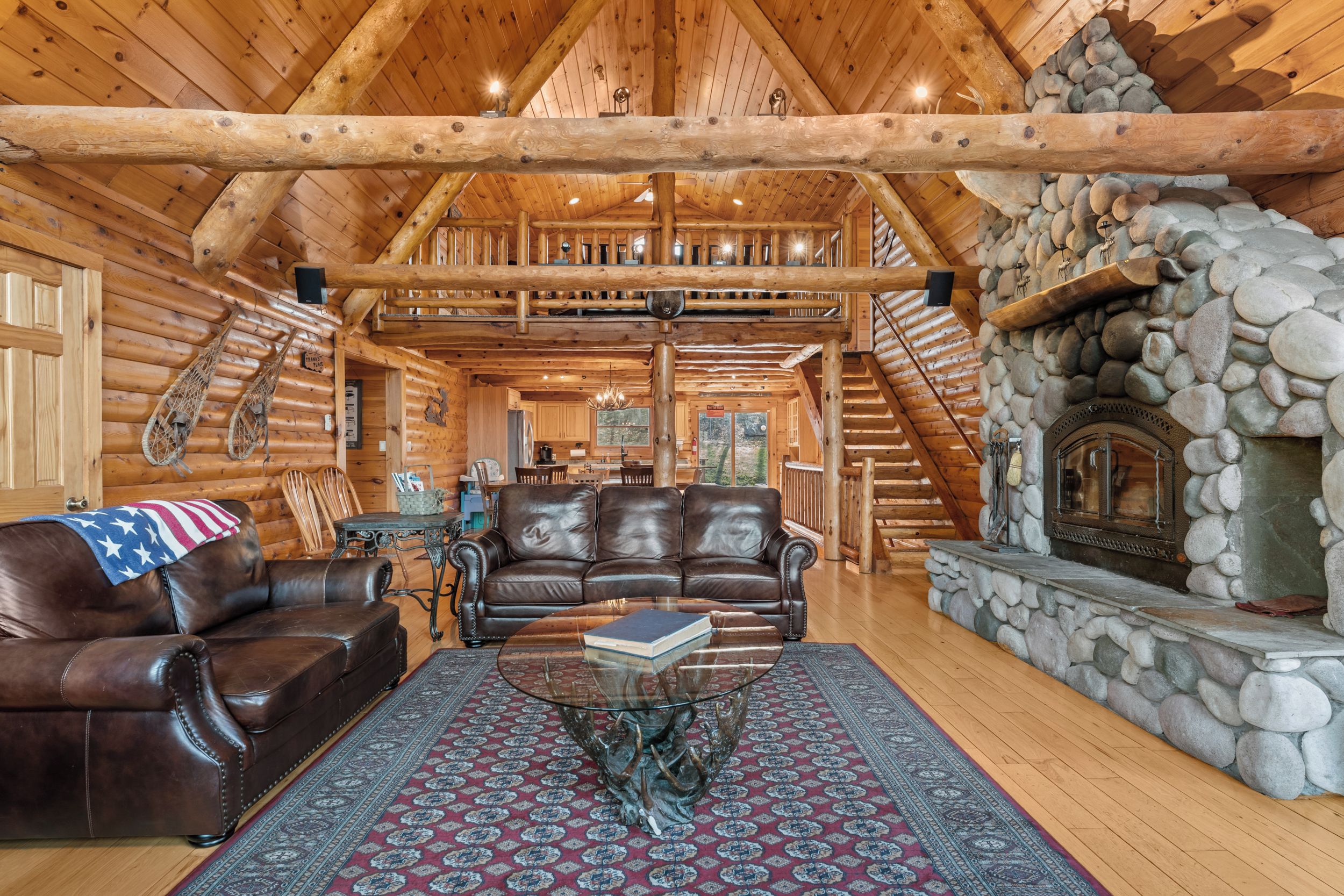 L’intérieur d’une cabane en rondins, avec une cheminée en pierre, des meubles en cuir et une vue sur l’extérieur.