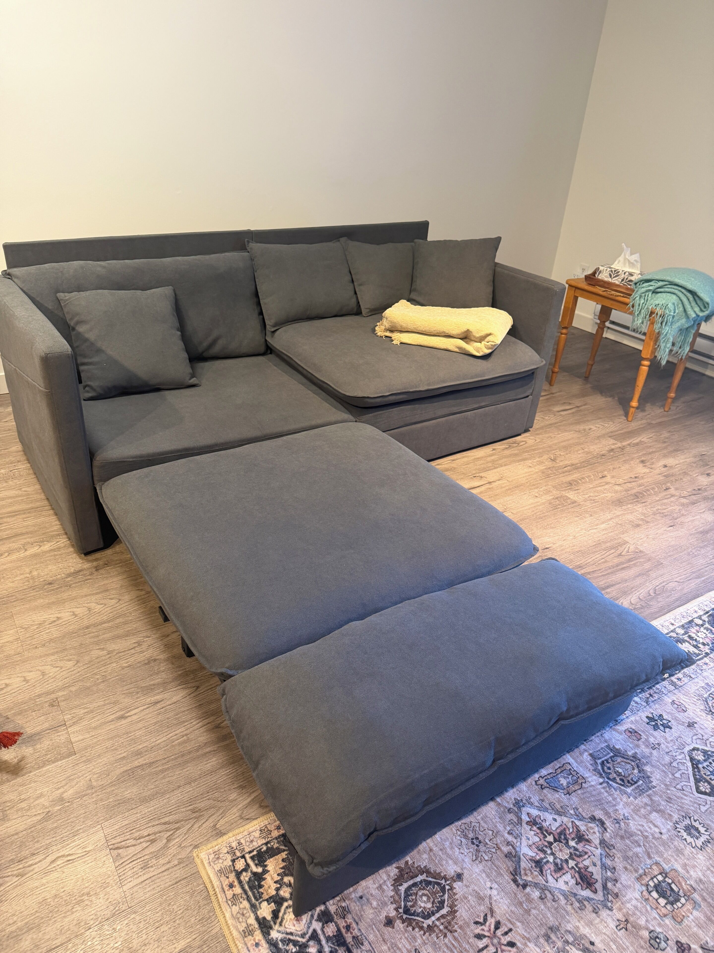 A grey sectional sofa with a matching ottoman, a small wooden side table, and a patterned rug on a wooden floor.