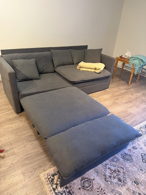 Un divano angolare grigio con poggiapiedi coordinato, un piccolo tavolino in legno e un tappeto decorato su un pavimento di legno.