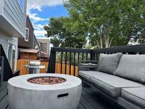 Eine Terrasse mit einer runden Feuerstelle, einem grauen Sofa und einem Holzzaun.