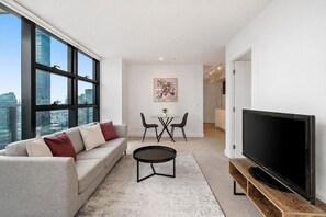 A modern living room with a sofa, a coffee table, and a TV. There is a dining area with a table and chairs. Large windows offer a view of the cityscape. - Sheraton Little Collins by Ready Set Stay (Melbourne)