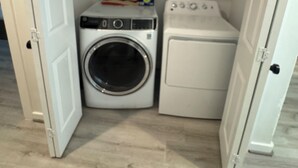 A laundry room with a washing machine, dryer, and shelves with laundry supplies. - 3BR home w/ AC, parking & W/D near Galleria/ Downtown (Houston)
