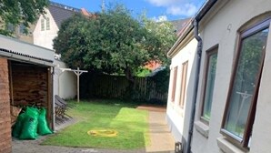 A paved backyard with a grassy lawn, a brick wall, and a white building with windows. - Comfortable and centrally located home in Odense (Odense)