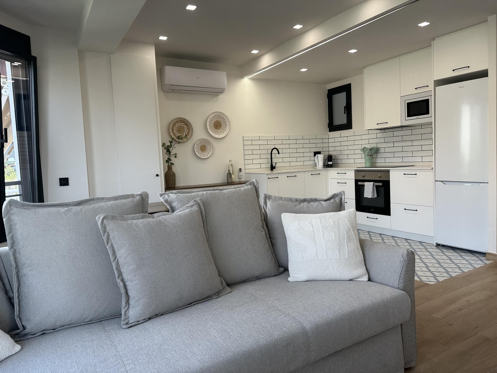 A modern living room with a grey sofa and matching cushions, a kitchen with white cabinets and a black oven, and a large window with curtains.
