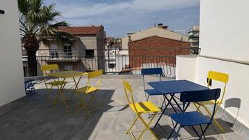 A terrace with yellow and blue foldable tables and chairs, a palm tree, and buildings in the background.