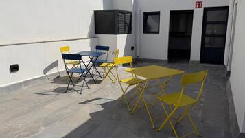 Un espace extérieur aménagé avec des chaises et des tables jaunes et bleues, un panneau indiquant la sortie de secours et l’entrée du bâtiment.