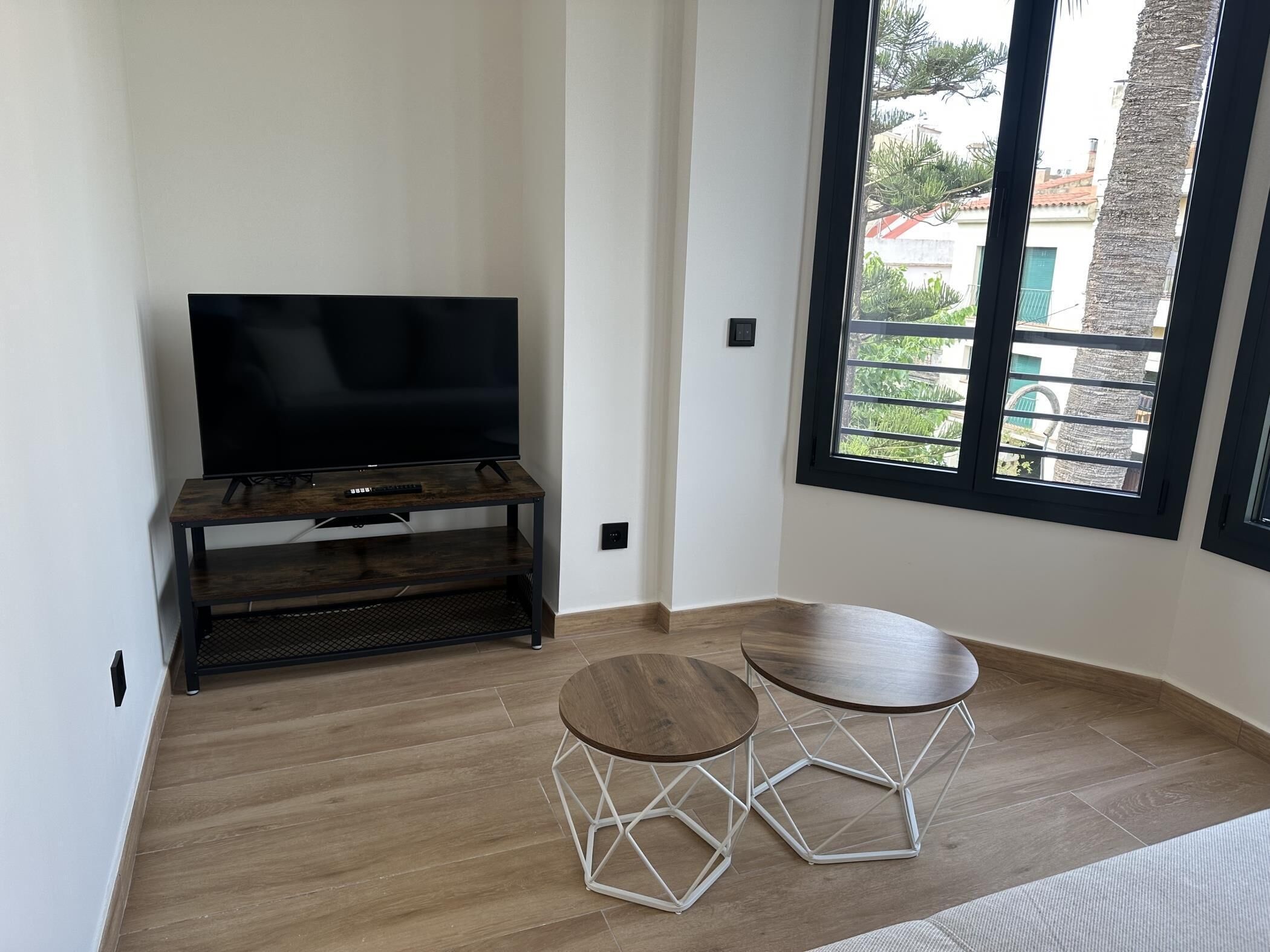 A room with a flat-screen TV on a wooden stand, a small table with a geometric base, and a large window with a view of buildings and trees.