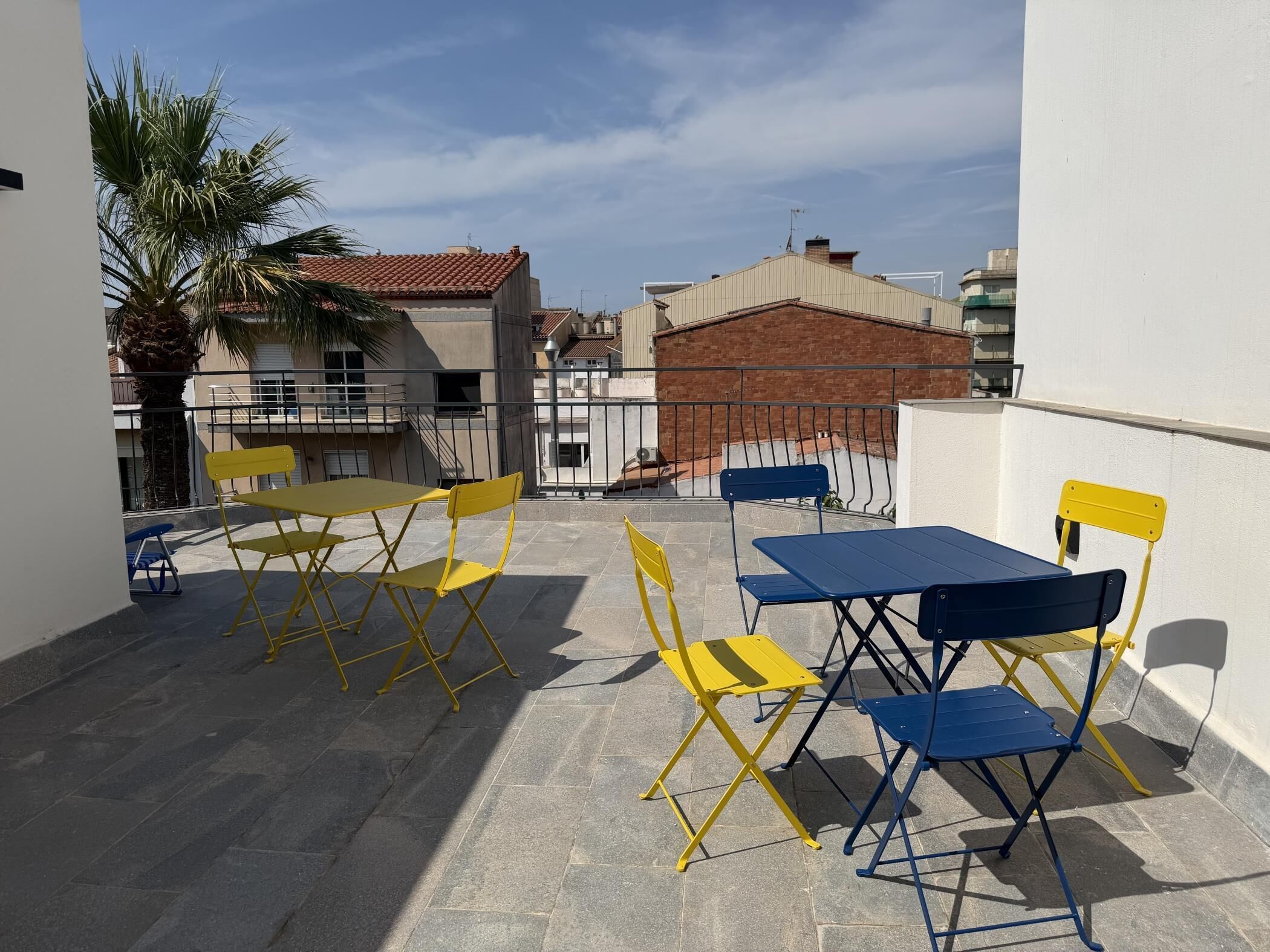 A terrace with yellow and blue foldable tables and chairs, a palm tree, and buildings in the background.