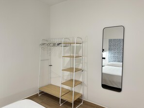 A room with a white metal shelf unit with wooden shelves, a white mattress, and a wall-mounted mirror reflecting a patterned wall. - Apartment with A/C and terrace only 250m beach (Calella)