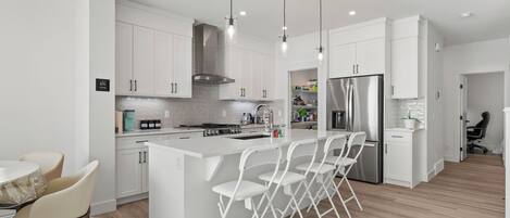A modern kitchen with white cabinetry, a central island with bar stools, a stainless steel refrigerator, and pendant lighting.