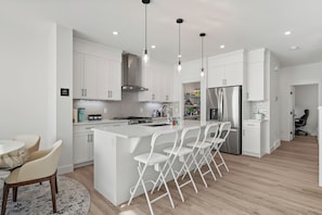 A modern kitchen with white cabinetry, a central island with bar stools, a stainless steel refrigerator, and pendant lighting. - Creekstone Retreat - South West Calgary - A Cozy Home for a Peaceful stay (Calgary)