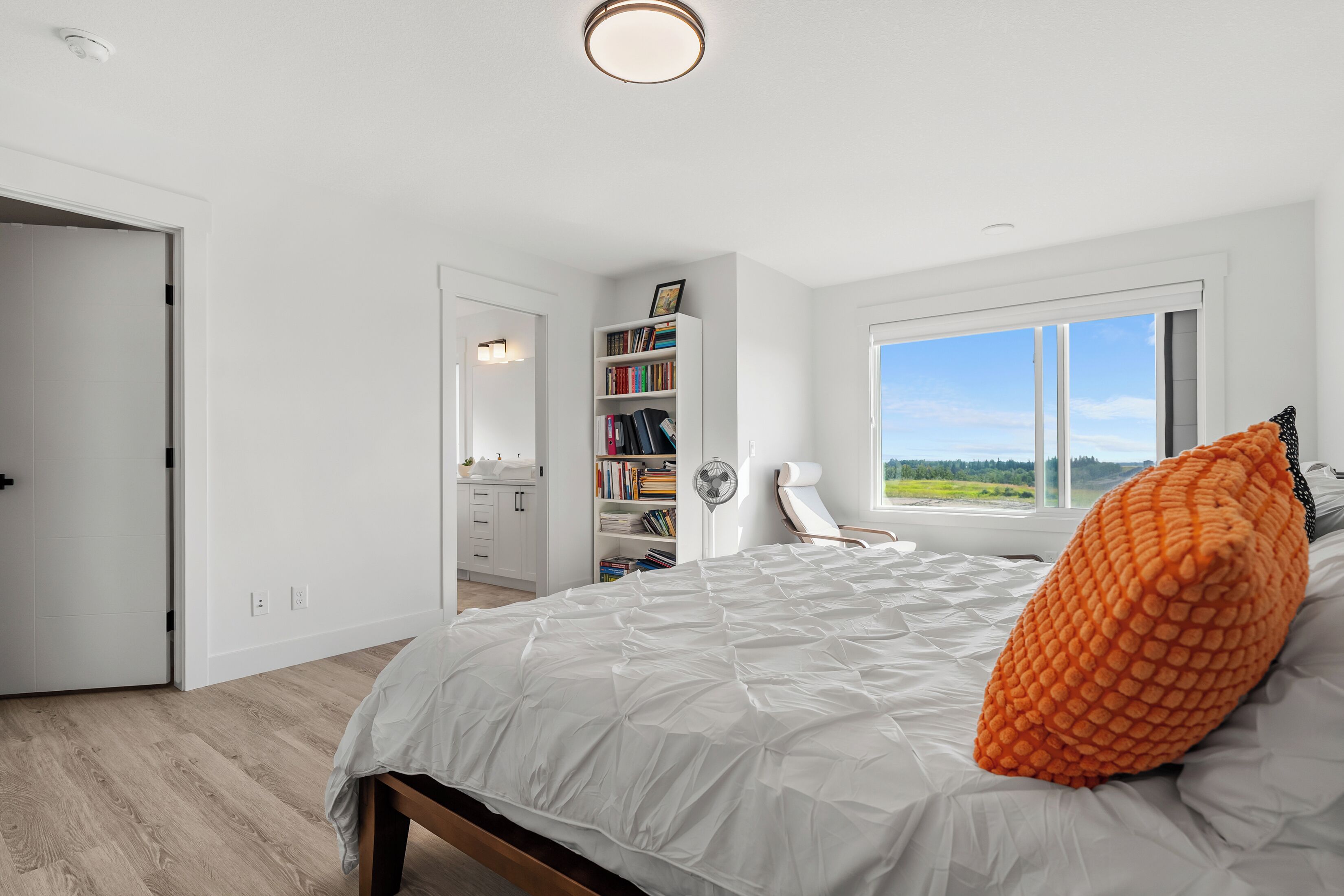 A bedroom with a large bed, a bookshelf, a chair, and a window with a view.