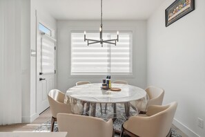 A dining area with a round table, six chairs, a chandelier, and a window with blinds. - Creekstone Retreat - South West Calgary - A Cozy Home for a Peaceful stay (Calgary)