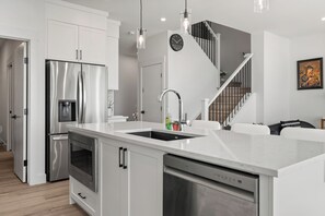 A modern kitchen with stainless steel appliances, a central island with a sink, and a staircase leading to an upper level. - Creekstone Retreat - South West Calgary - A Cozy Home for a Peaceful stay (Calgary)