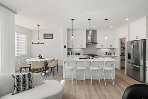 A modern kitchen with white cabinetry, a stainless steel refrigerator, and a dining area with a round table and chairs. - Creekstone Retreat - South West Calgary - A Cozy Home for a Peaceful stay (Calgary)