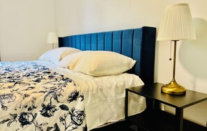 Una cama con cabecera azul, almohadas blancas y colcha estampada con motivos florales. Se observa una mesita de noche con una lámpara.