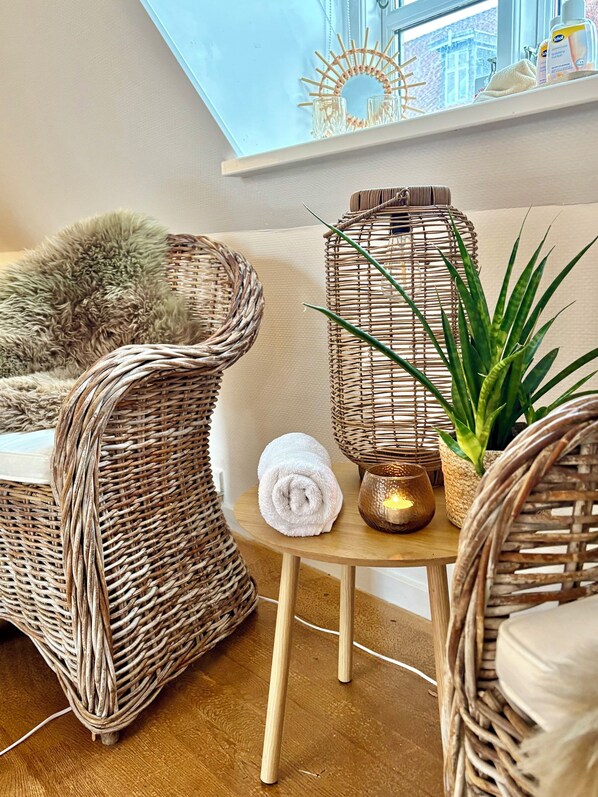 A wicker chair, a small table with a candle, and a potted plant in a room with a window and a view of a building.
