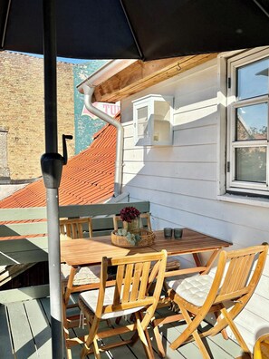 An outdoor patio with a table and chairs, a black umbrella, and a view of a building with a window and a door. - Holiday apartment in heart of Møn (Stege)