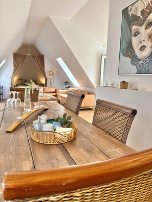 A loft-style room with a high ceiling, a dining table with wicker chairs, a hanging light fixture, and a painting on the wall.