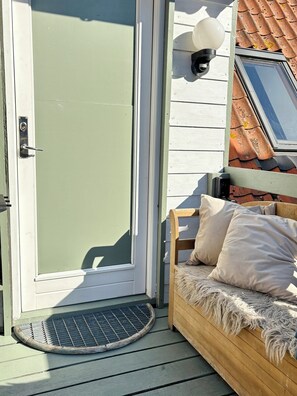 A door with a metal handle, a small porch area with a wooden bench and cushions, and a wall-mounted light fixture.