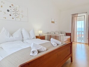 A hotel room with a large bed, a wooden headboard, a nightstand, a lamp, a crib, and a view of the outdoors through a glass door. - Apartment Stina by Interhome (Novi Vinodolski)