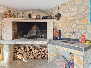 A stone fireplace with a metal grate, a stone wall, a sink, and a pile of firewood. - Apartment Loza by Interhome (Novi Vinodolski)