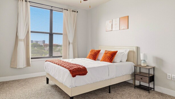 A bedroom with a bed, a nightstand, a ceiling fan, and a window with curtains. - Downtown Loft w/ Pool, Gym & Parking - Walk to Sundance Square (Fort Worth)