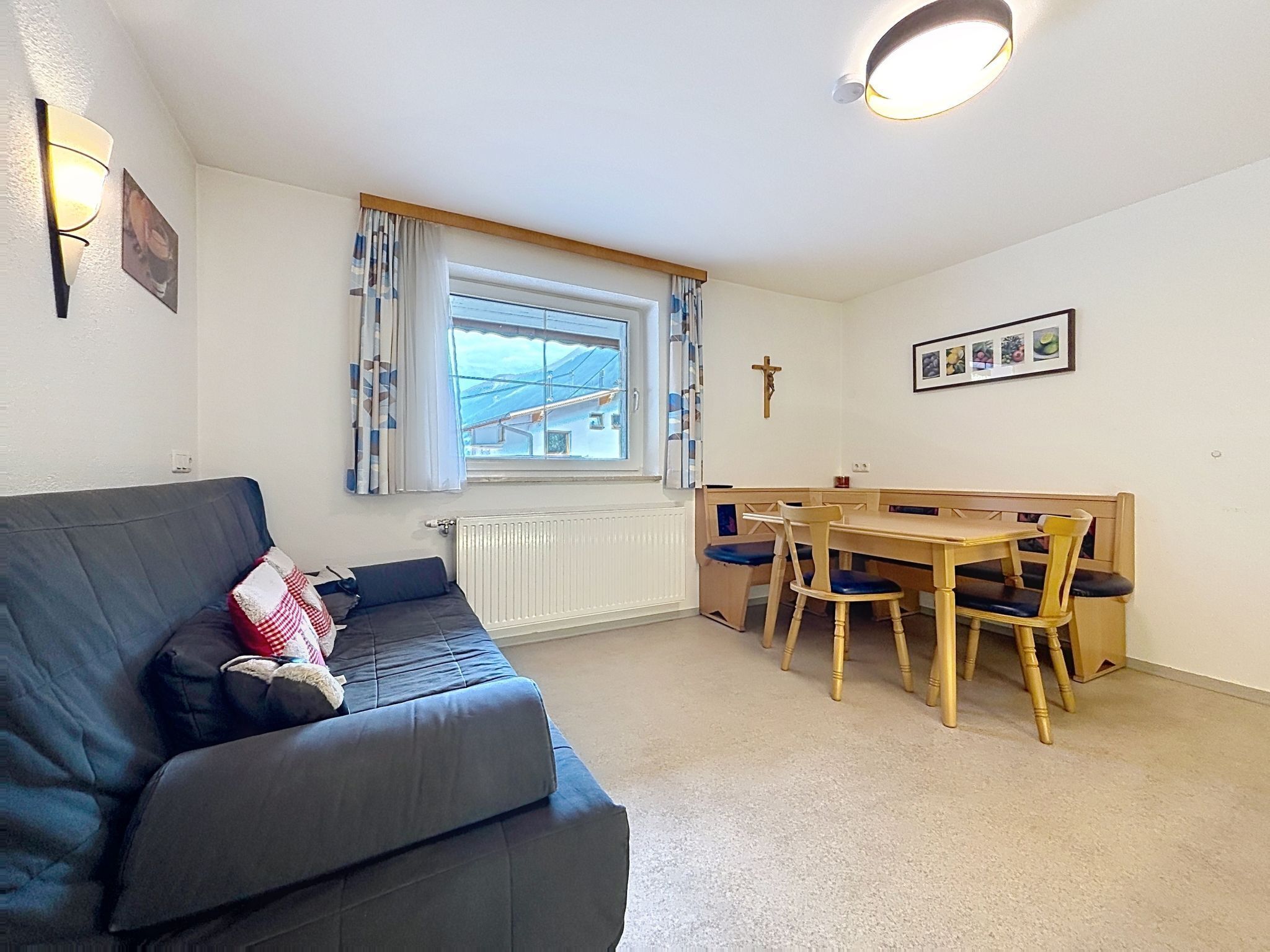 A room with a sofa, a dining table with chairs, a window with curtains, a radiator, and a wall-mounted cross.