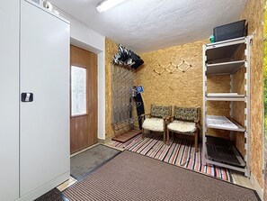 Ein kompakter Raum mit Holztür, Kleiderhaken mit aufgehängten Kleidungsstücken, zwei gemusterte Stühle, ein kleiner Tritt zur Tür und ein Regalelement mit einem gefalteten Gegenstand darauf.