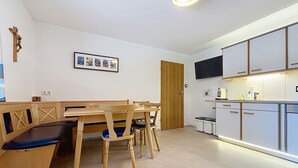 Una habitació amb taula i cadires de fusta, una petita cuina amb armaris i un televisor, i una porta.