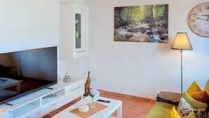 A living room with a flat-screen TV, a coffee table with decorative items, a sofa with cushions, a painting of a forest stream, and a wall clock. - Das Dorfnest by Interhome (Wanna)