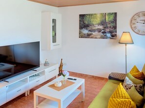 Ein Wohnzimmer mit Flachbildfernseher, Couchtisch mit Dekoration, einer Couch mit Kissen und einem Wandbild eines Waldflusses. An der Wand hängt eine Uhr.