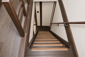 Eine Treppe mit Holzdielen und Metallgeländern, die zu einer Zwischenebene mit einer hellen Wand und einem kleinen schwarzen Lüftungsgitter führt.