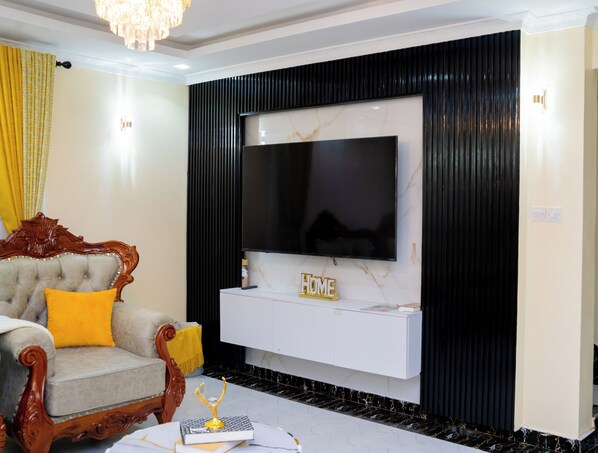 A living room with a television mounted on a white cabinet, a plush armchair with a yellow cushion, and a decorative golden object on the coffee table.