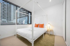 Un dormitorio moderno con cama con dosel, una mesita de noche con lámpara y vistas a un paisaje urbano a través de amplios ventanales.