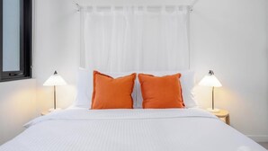 Ein Bett mit weißer Bettwäsche und zwei orangenen Kissen, daneben zwei Nachttische aus Holz mit Nachttischlampen.