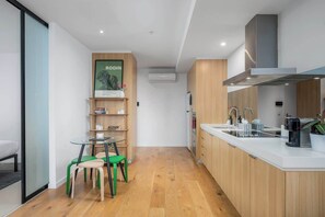 A modern kitchen with a white countertop, wooden cabinets, and a built-in stove. There is a small dining area with a glass table and green chairs. - LaneStay - 2BR Southern Cross & Marvel, Pool & Gym (Melbourne)