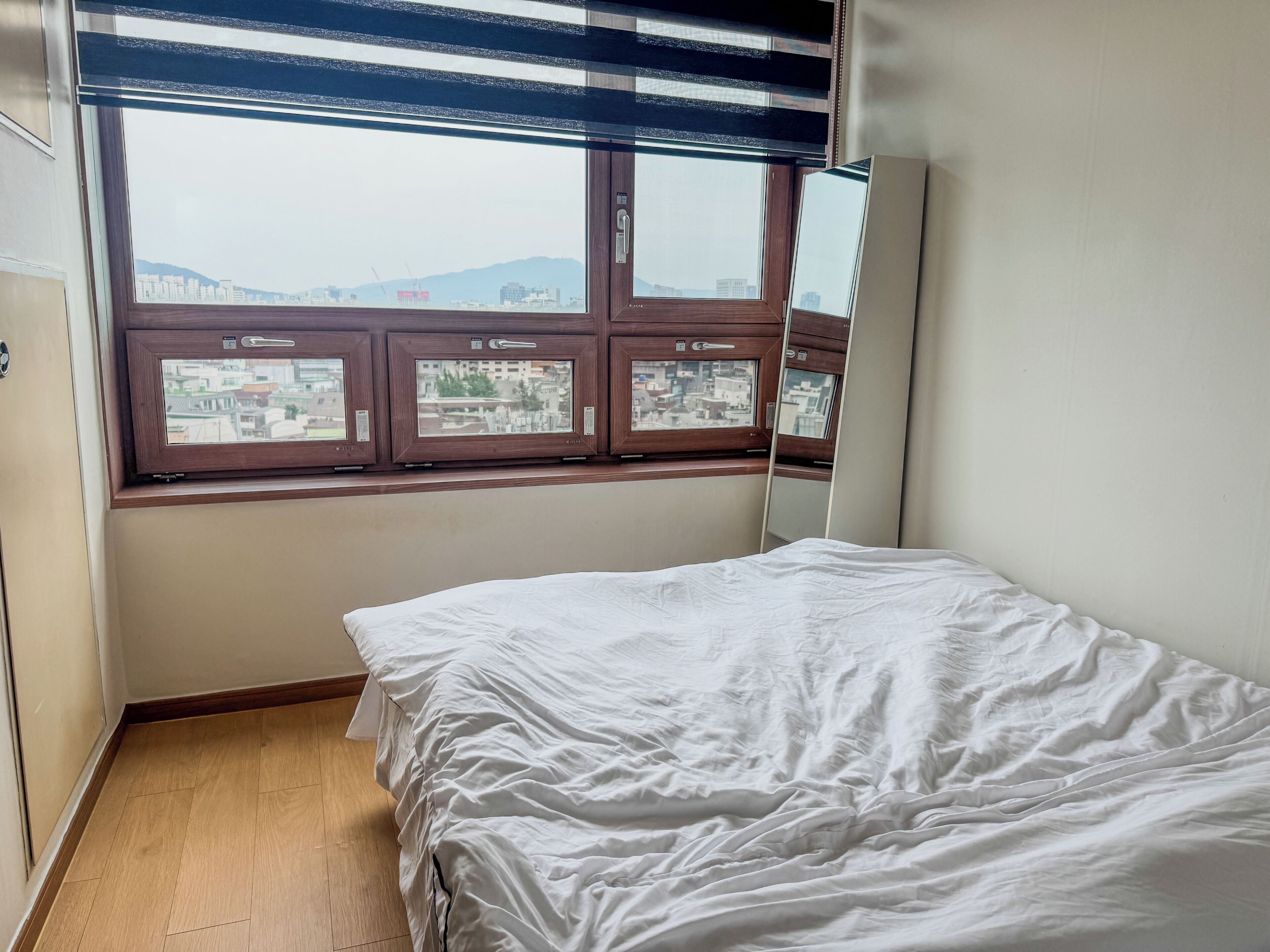 A room with a bed, a window with blinds, and a view of a cityscape.